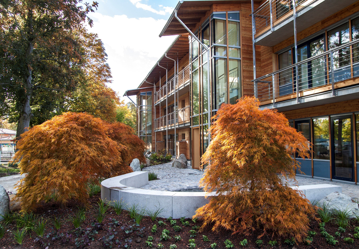 Unser Firmengebäude mit großen Fensterfronten und Balkonen, davor ein gestalteter Außenbereich mit orangefarbenen Bäumen im Herbst.